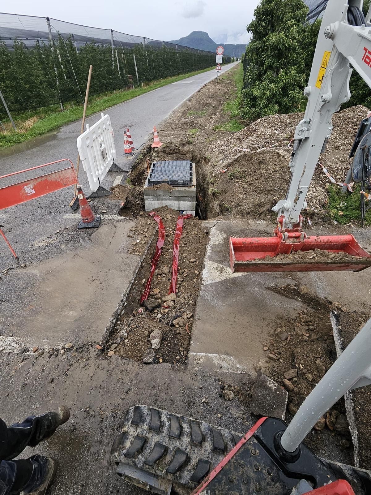Trincea lungo una strada di campagna con pozzetto in cemento posato, coni e barriere di cantiere